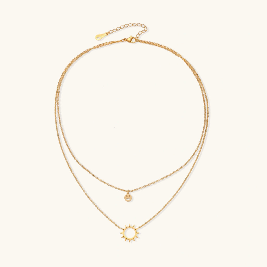 Gold layered necklace set with sunburst and smiley face pendants on delicate chains against white background.